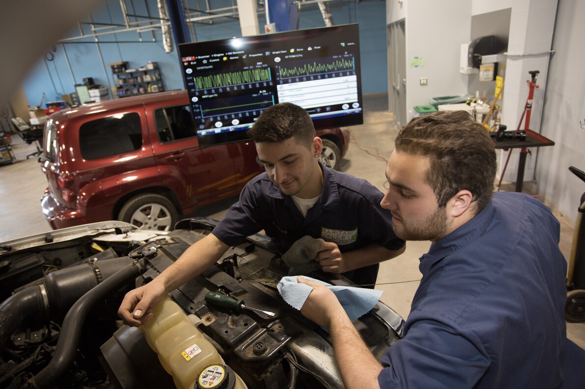 2 male students working on car with red car in background