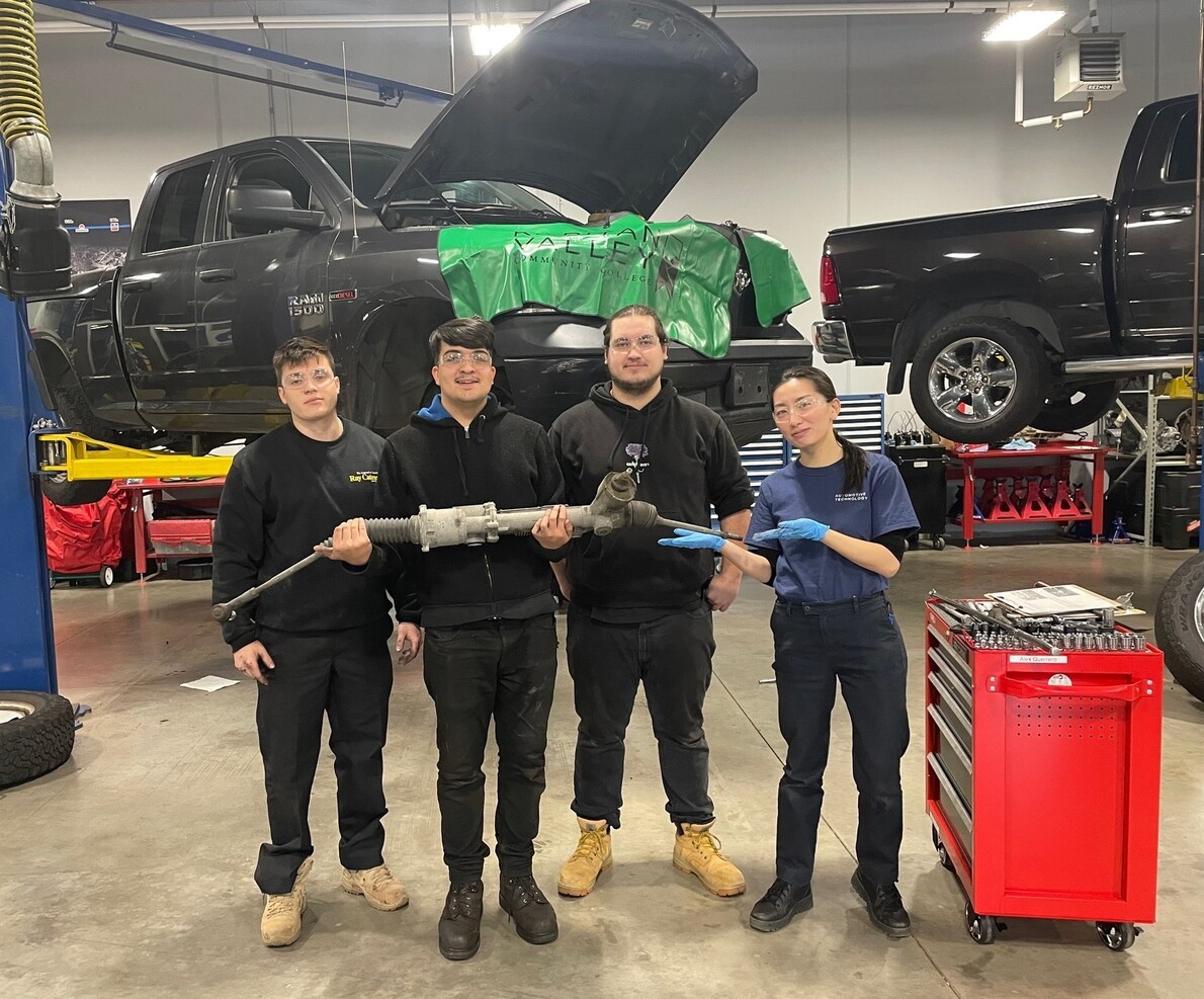 3 male and 1 female automotive students in front of lifted cars