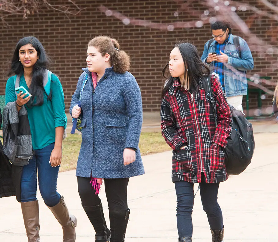 3 girls in coats walking outside with male student on phone