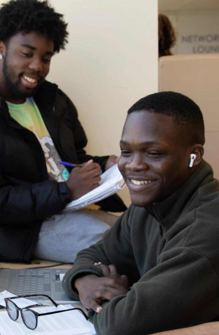 2 male students smiling, working on laptop, writing