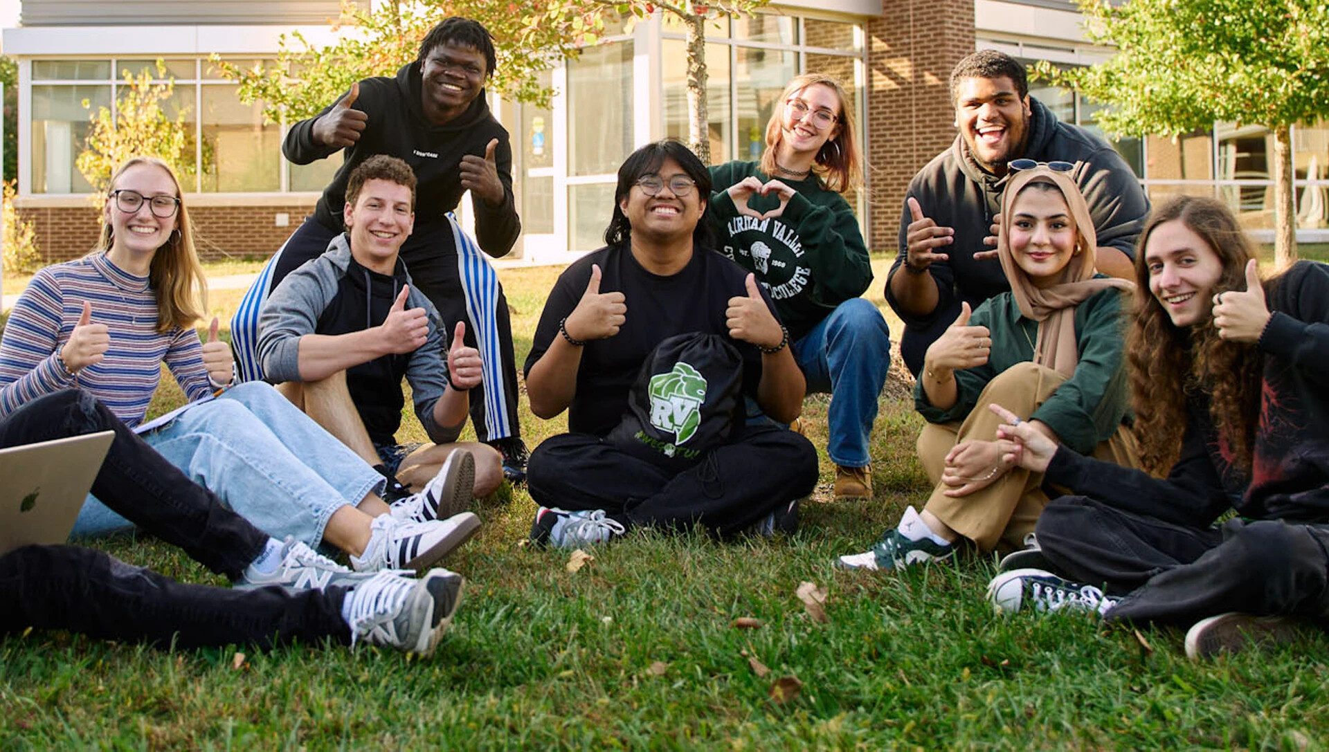 large group of college students sitting in the grass, a few giving a thumbs up