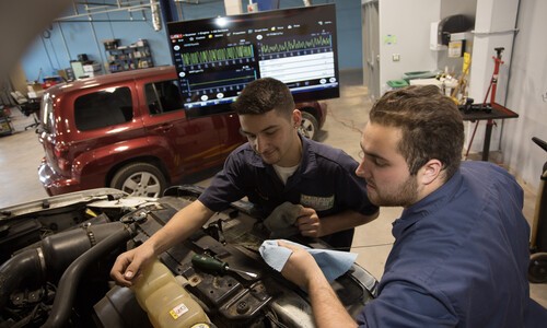 2 male students working on car with red car in background