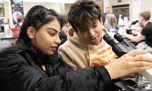 female student in black jacket with male student looking at microscope