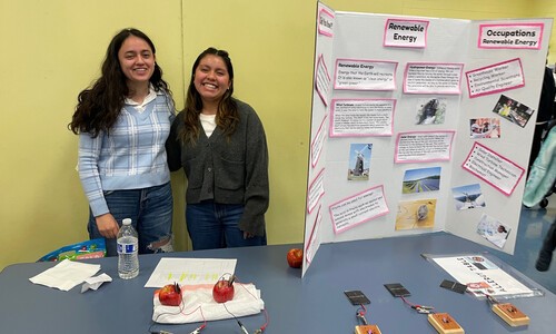 2 female students standing next to poster board on table