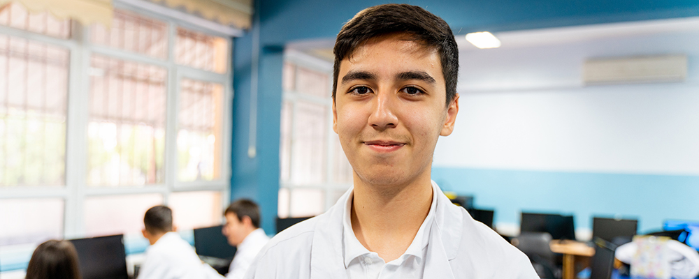 A pharmacy technician student wearing a white lab coat stands in a classroom computer lab, smiling at the camera while other students work in the background.