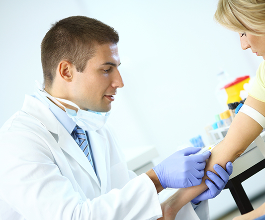 A phlebotomy assistant wearing gloves and a lab coat draws blood from a patient’s arm in a clinical setting.