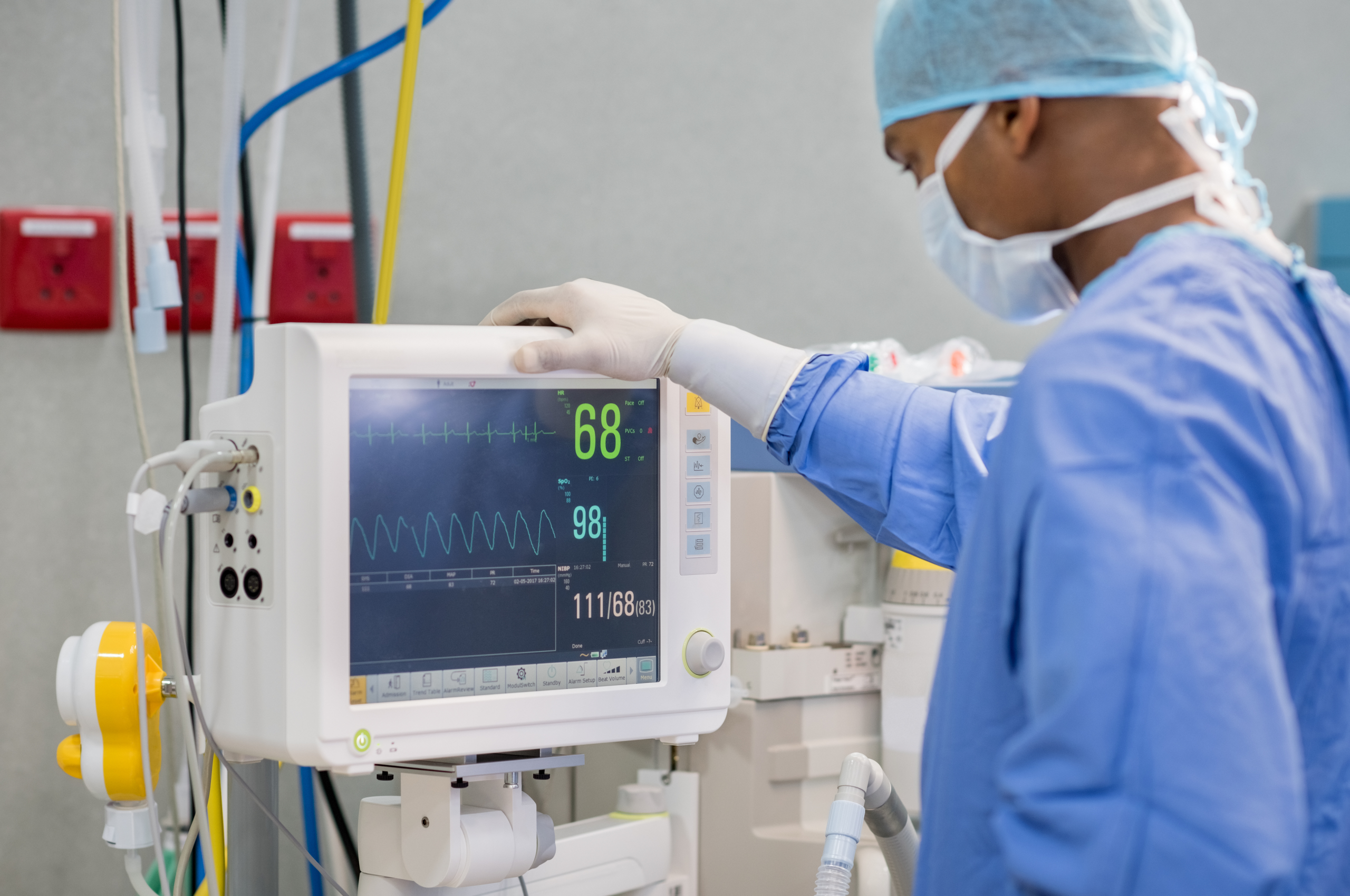 A healthcare worker wearing surgical scrubs, gloves, a mask, and a cap adjusts a patient monitoring machine in a clinical setting. The monitor displays vital signs including heart rate, oxygen saturation, and blood pressure readings.