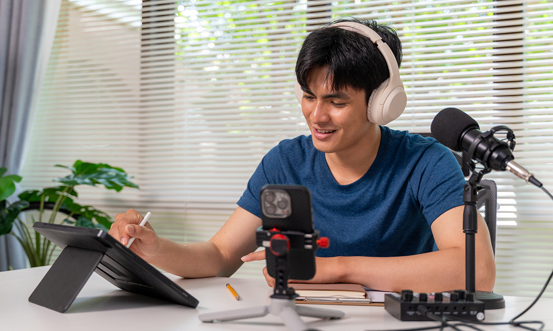 Young Asian man recording a podcast at home using a tablet, headphones, and microphone, sitting at a desk in a modern workspace with audio equipment.