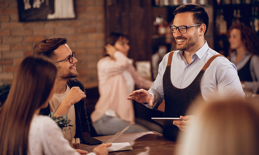 Smiling waiter holding digital tablet while taking order from a couple in a bar.