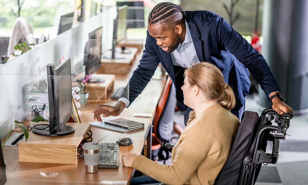Black businessman assisting a female coworker in a wheelchair as they engage in productive collaboration within a modern open office. The scene conveys a sense of support and teamwork in the workplace environment.