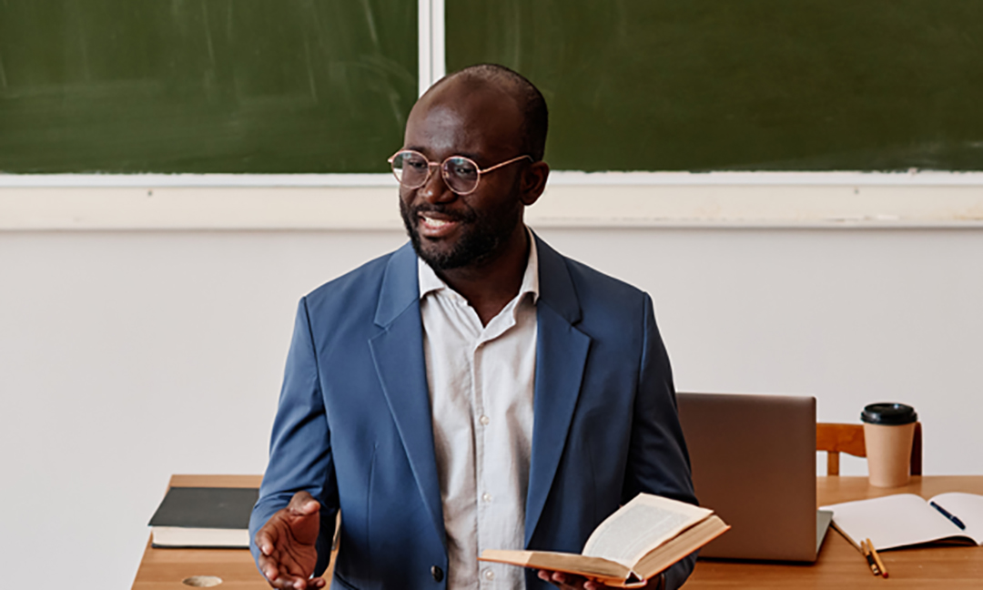 Teacher in a suit in front of a chalk board, with open book in his hand.