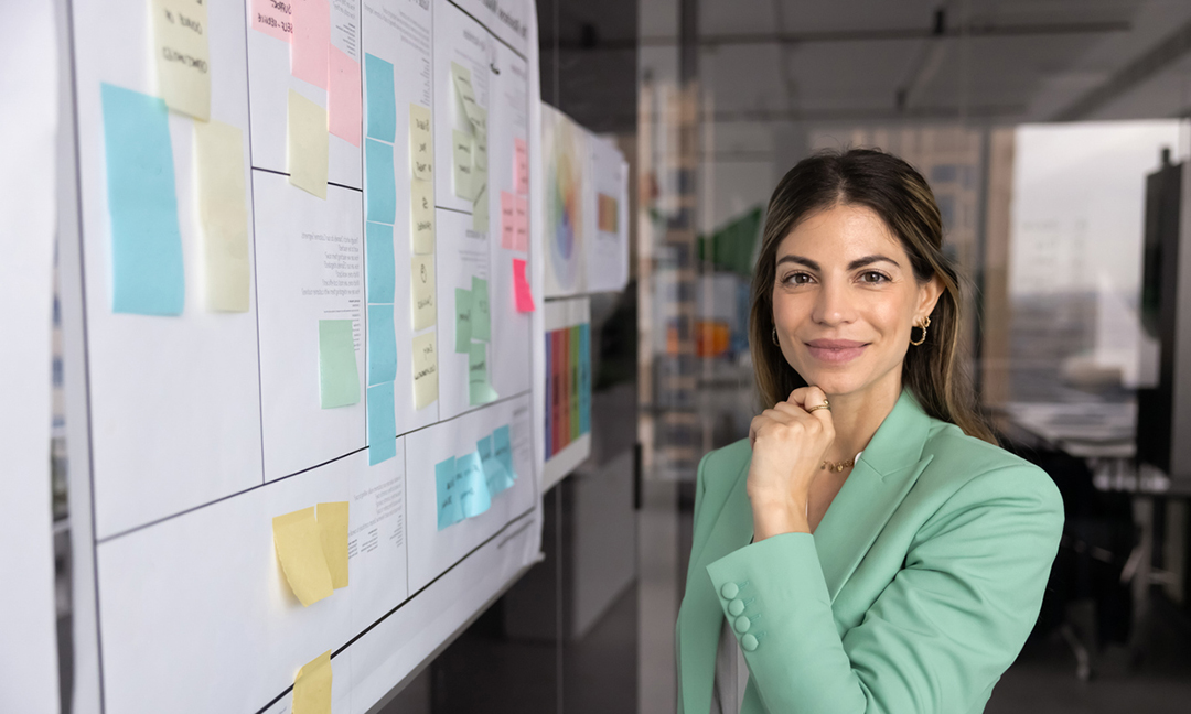 Portrait of attractive Hispanic businesswoman, office employee stands thoughtfully in front of large planning board filled with sticky notes and documents, analyzing or evaluating strategy and ideas
